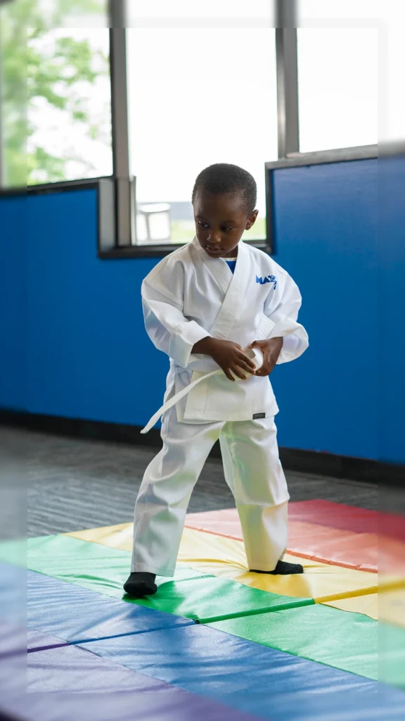 Young child in white martial arts uniform practicing stance on colorful mat.