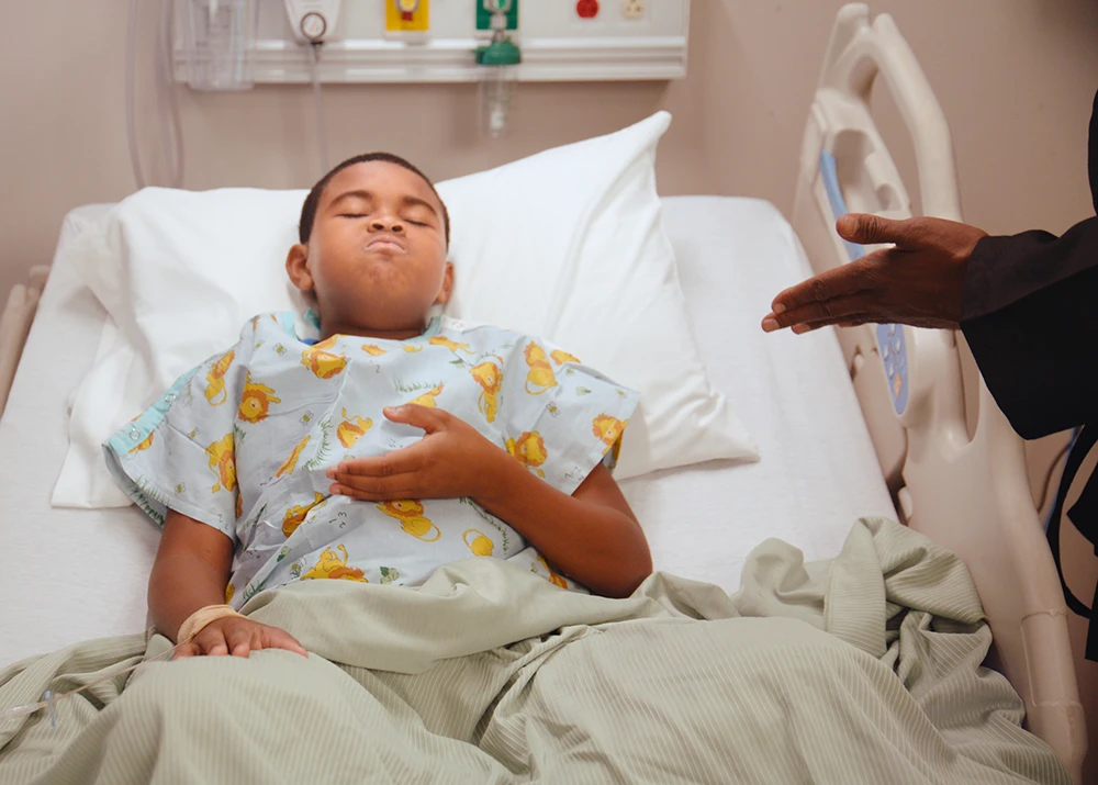 Child in hospital bed wearing gown with hand raised in breathing gesture.